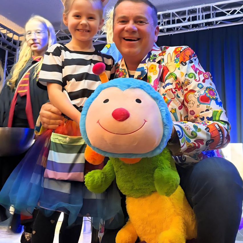 Ein Mann in einem bunten Anzug hält ein großes Plüsch-Affenspielzeug, während ein junges Mädchen in einem schwarz-weiß gestreiften Shirt lächelt. Dahinter hält eine Frau in einem Kostüm eine Tasche. Die Umgebung scheint eine Bühne oder ein Event zu sein.