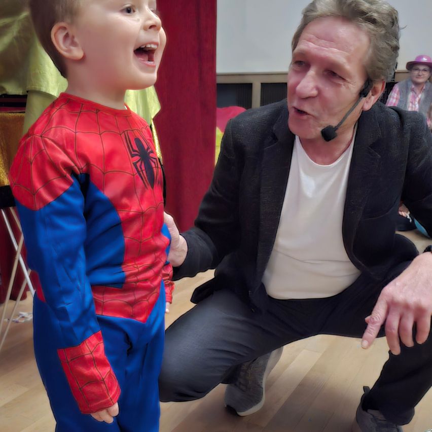 Ein junger Junge in einem Spider-Man-Kostüm lacht, während ein Mann mit einem Mikrofon neben ihm hockt. Dahinter sitzt eine Person auf einem Stuhl, und ein roter Vorhang hängt im Hintergrund.