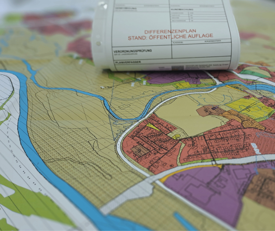 Eine aufgerollte Karte mit einem 'Differenzplan'-Etikett darauf liegt auf einer anderen Karte. Die Karte darunter zeigt eine Stadt mit verschiedenfarbigen Bereichen. Es gibt einen Fluss, der durch die Stadt fließt.