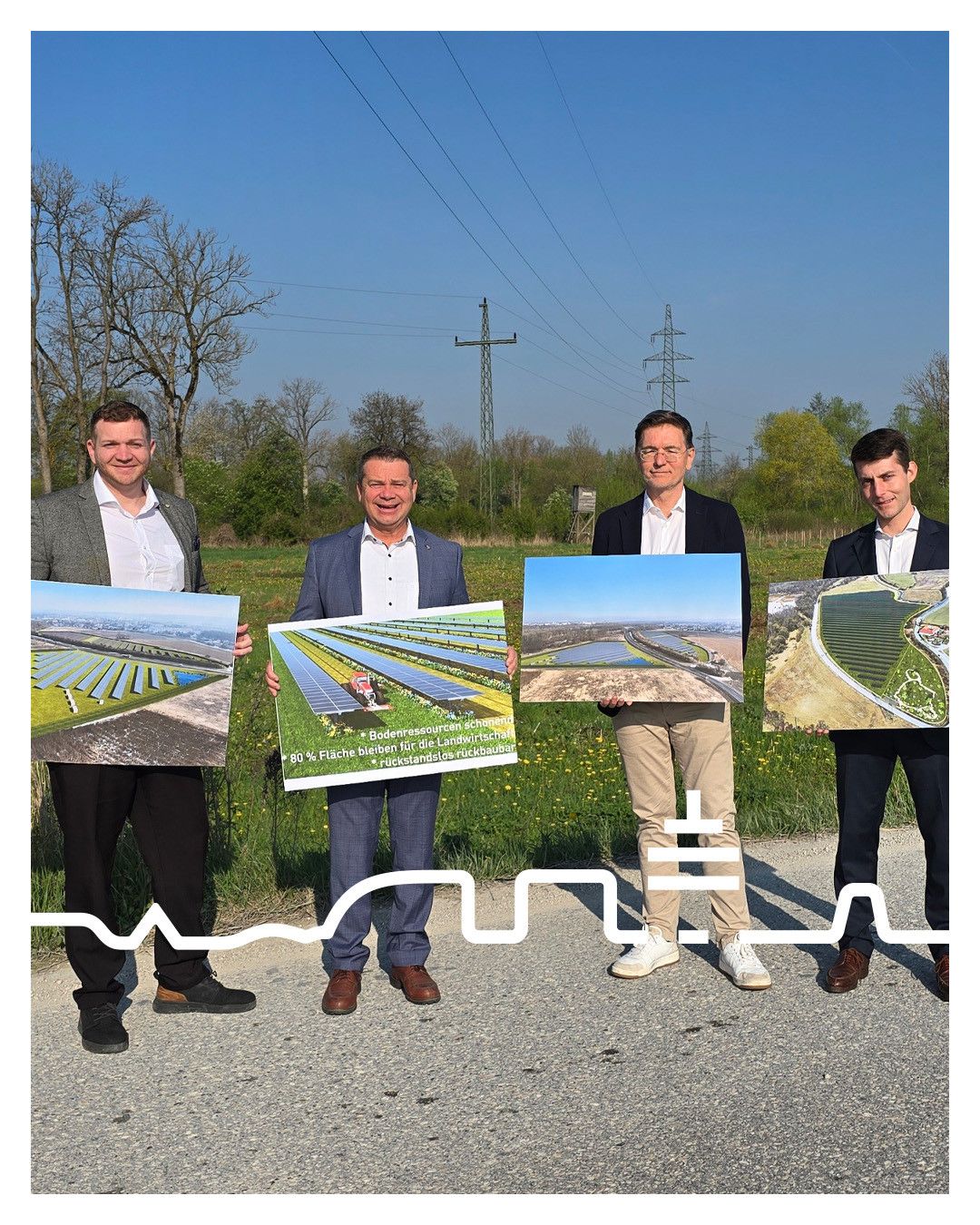 Vier Männer stehen auf einer Straße und halten große Plakate von Solarparks hoch. Sie sind geschäftlich gekleidet, wobei der Mann in der Mitte ein Plakat mit einem Traktorbild hält. Im Hintergrund sind Felder und Stromleitungen zu sehen.
