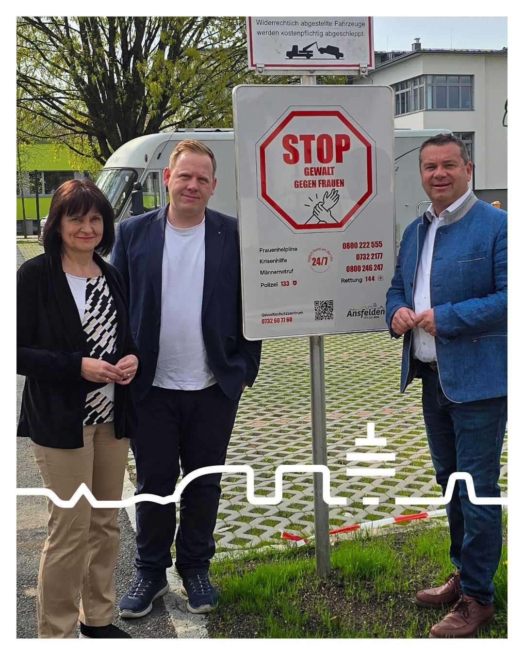 Drei Personen stehen neben einem STOP-Schild mit einer Botschaft gegen Gewalt an Frauen. Eine Frau und ein Mann tragen formelle Kleidung und stehen links und rechts.