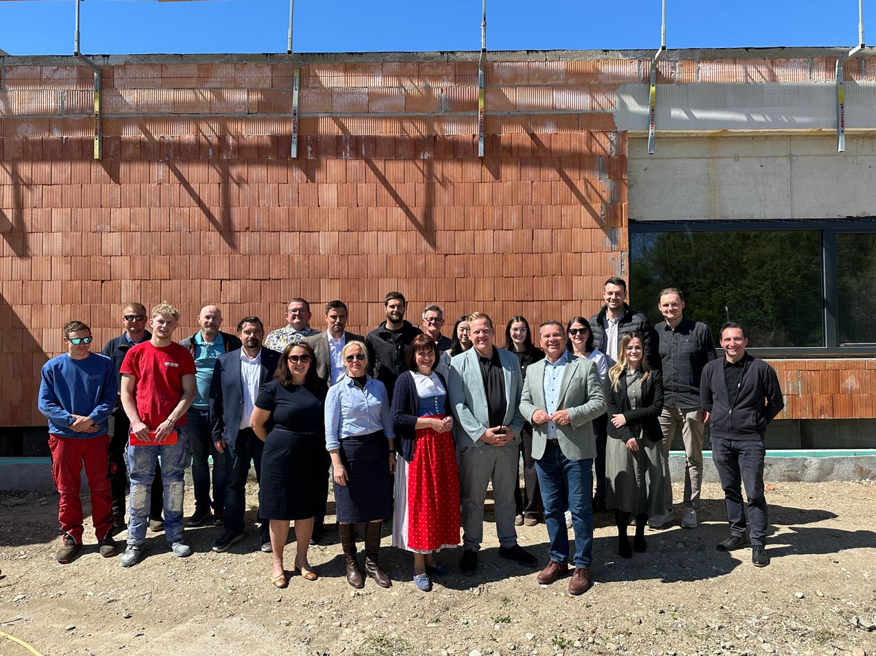 Eine Gruppe von Menschen steht vor einem im Bau befindlichen Gebäude. Sie sind in verschiedenen Outfits gekleidet, einige mit Sonnenbrillen, und scheinen für ein Foto zu posieren.