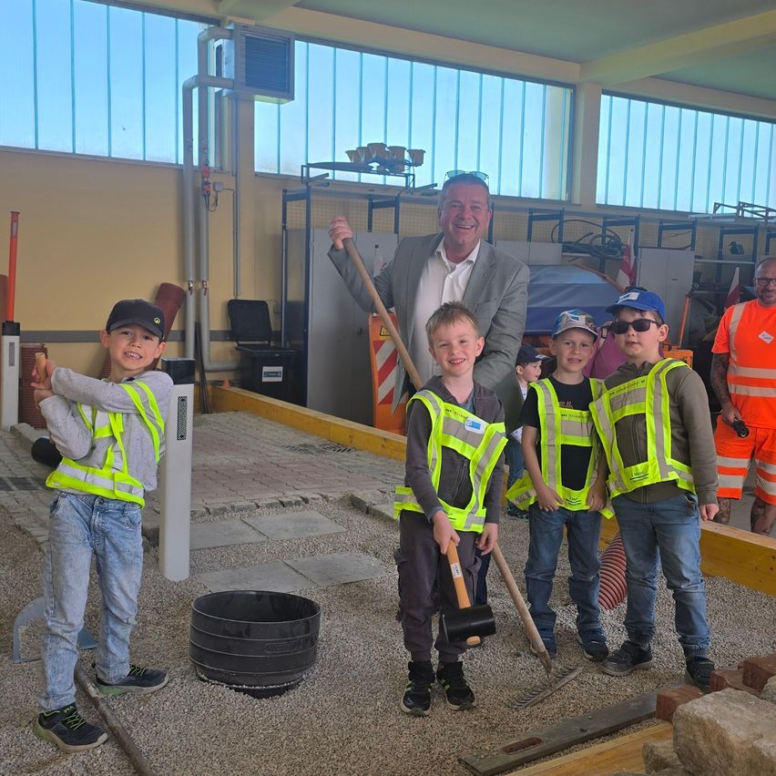 Vier Jungen und ein Mann im Anzug posieren für ein Foto. Sie tragen Warnwesten und halten Bauwerkzeuge. Auf dem Boden liegt ein schwarzer Gegenstand.