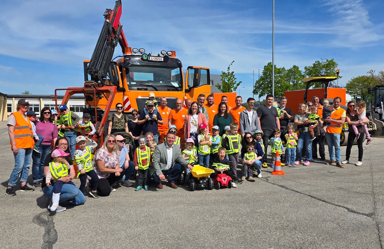 Eine Gruppe von Erwachsenen und Kindern in Warnwesten posiert mit einem Baufahrzeug auf einem Parkplatz an einem sonnigen Tag.