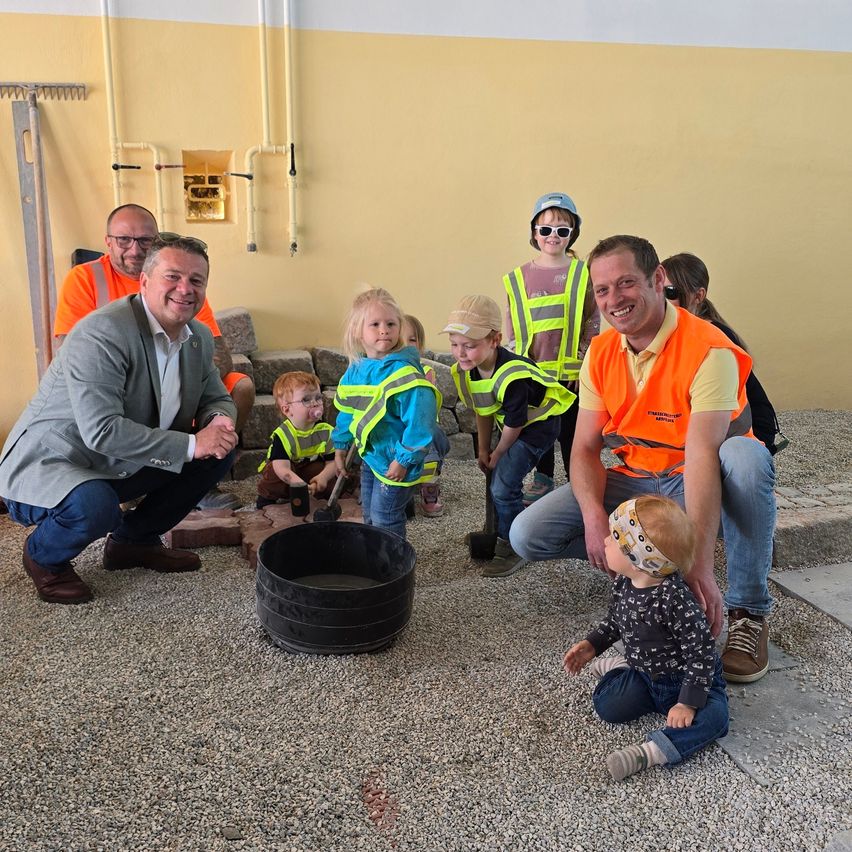 Eine Gruppe von Erwachsenen und Kindern versammelt sich um einen Zementkübel, einige tragen Sicherheitswesten und Helme, in einem Baubereich.