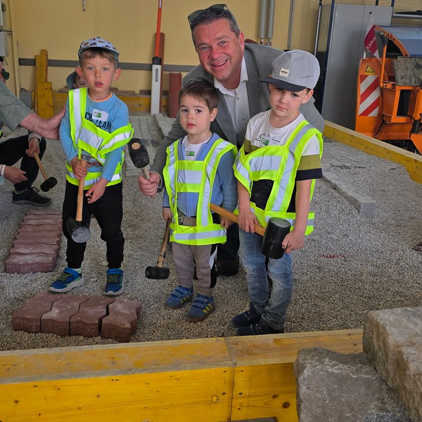 Drei Jungen in Bauwesten halten Hämmer in der Hand und posieren mit einem Erwachsenen Mann. Sie befinden sich auf einer Baustelle. Hinter ihnen ist ein Baufahrzeug geparkt.