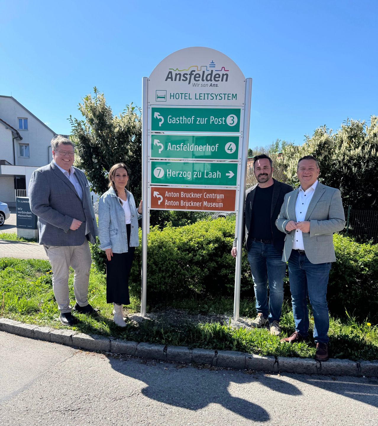 Vier Personen posieren vor einem Schild für Ansfelder, einem Hotelsystem mit Wegbeschreibungen zu verschiedenen Zielen.