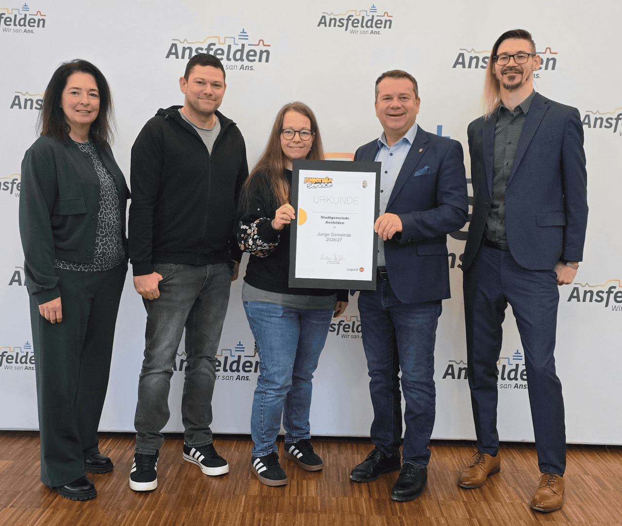 Fünf Personen stehen gemeinsam auf einem Holzboden, eine Frau links und ein Mann rechts, beide lächeln. Die Frau hält ein Zertifikat, und der Mann hält ein Zertifikat und eine Mappe. Hinter ihnen zeigt eine weiße Hintergrundwand den Text 'Ansfelder' und 'Junge Gemeinde Ansfelder'.