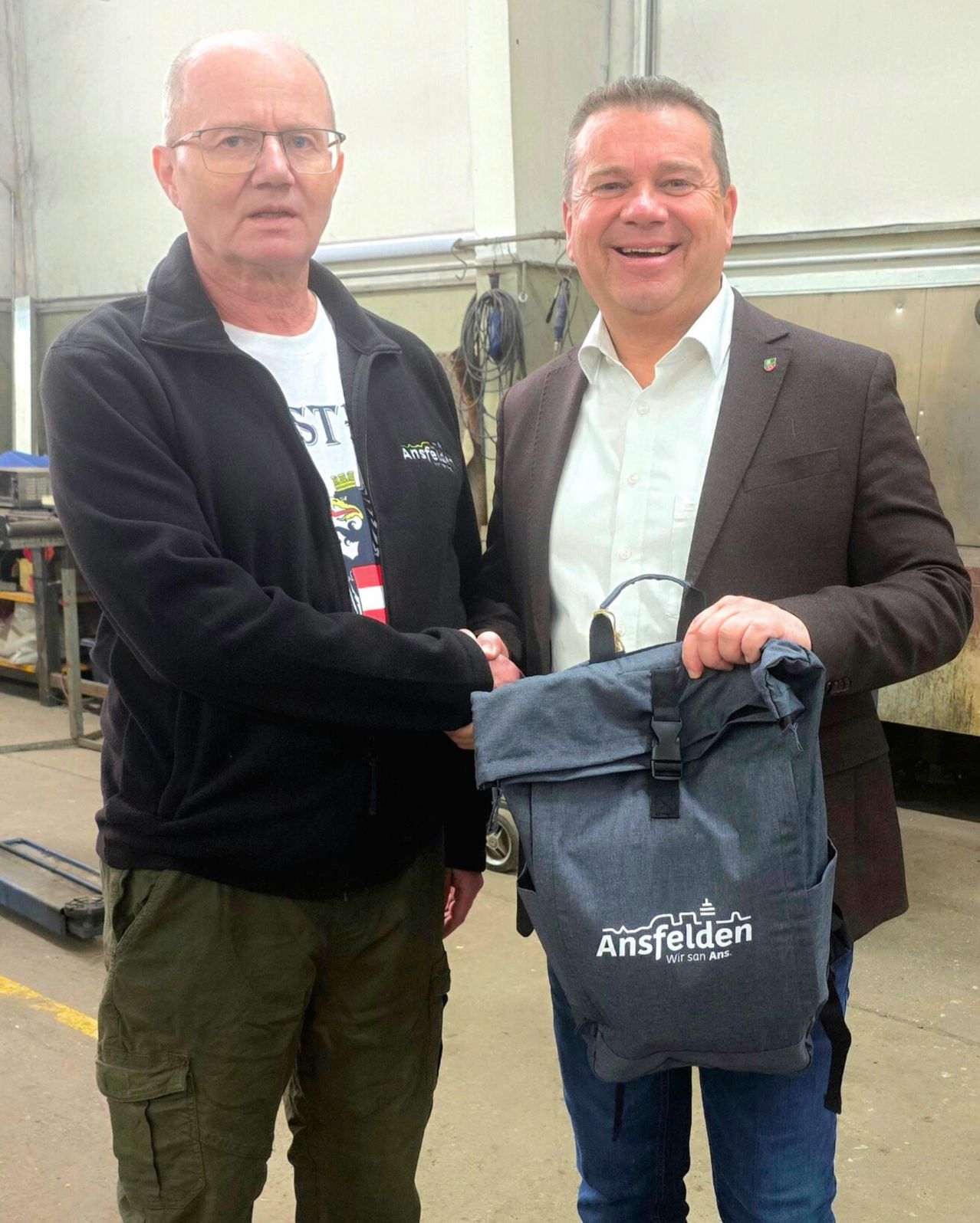 Zwei Männer stehen in einer industriellen Umgebung, einer hält eine Tasche, während der andere seine Hand zum Händeschütteln ausgestreckt hat. Der Mann auf der linken Seite trägt eine Brille und eine schwarze Jacke, während der Mann auf der rechten Seite einen Anzug trägt.