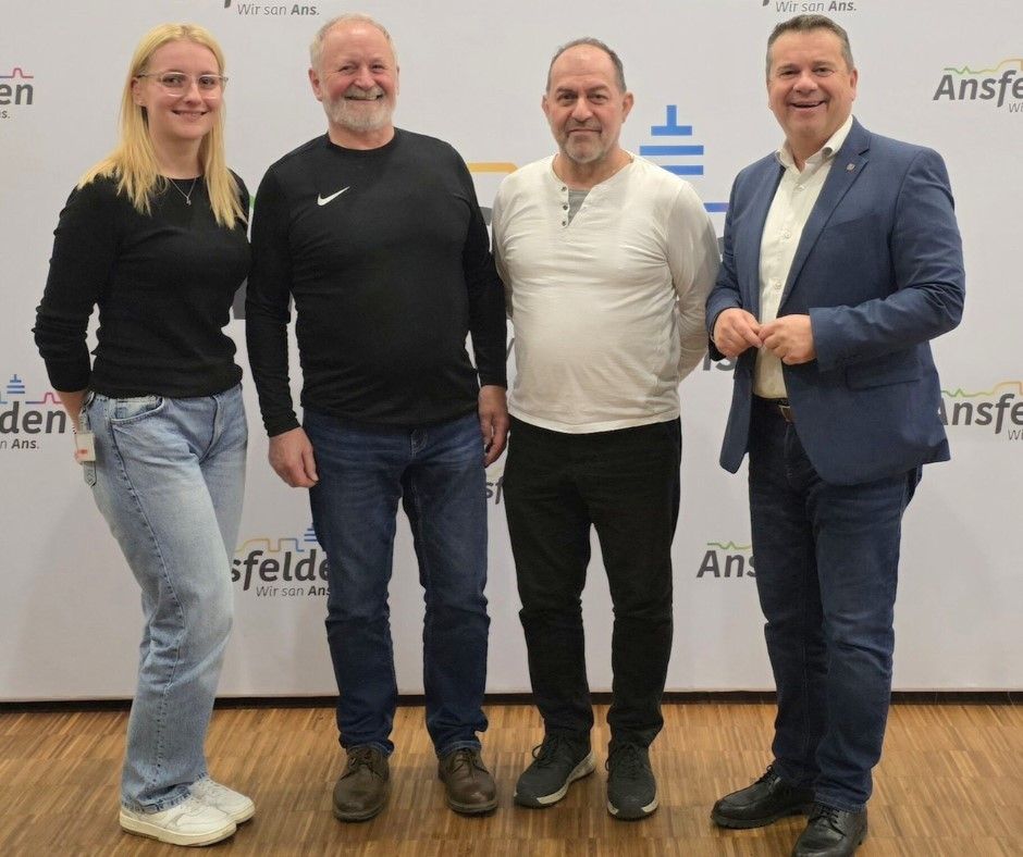 Vier Personen stehen vor einem weißen Hintergrund. Drei Männer und eine Frau lächeln. Der Mann links trägt ein schwarzes Langarmhemd und blaue Jeans. Der Mann in der Mitte trägt ein weißes Hemd und schwarze Hose. Der Mann rechts trägt einen blauen Anzug. Die Frau links trägt ein schwarzes Hemd und Jeans.