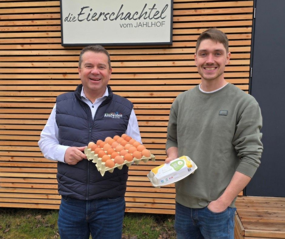 Zwei Männer stehen vor einer Holzwand. Einer hält ein Eiertablett und der andere eine Eierschachtel. Hinter ihnen steht ein Schild mit der Aufschrift 'die Eierschachtel vom JAHLHOF.'