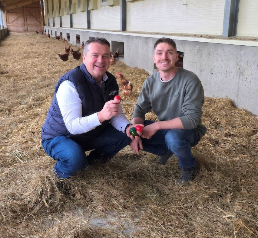 Zwei Männer sitzen auf Heu in einem Geflügelstall, lächeln und halten farbige Eier. Hühner laufen im Hintergrund herum.