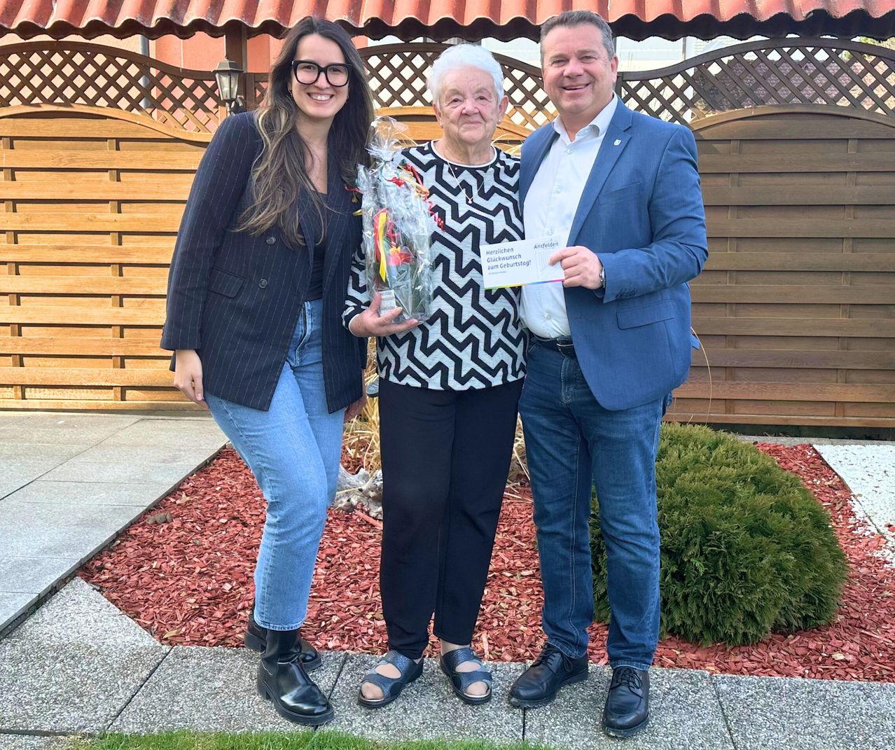 Drei Menschen posieren für ein Foto, eine Frau hält einen Blumenstrauß und ein Mann ein Zertifikat. Sie alle lächeln. Die Frau trägt eine Brille und Stiefel. Der Mann trägt einen Anzug. Dahinter befindet sich ein Holzzaun.