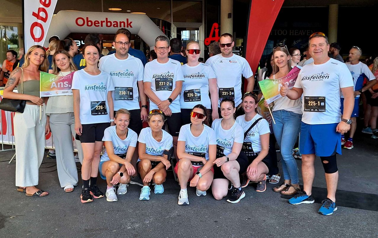 Eine Gruppe von Menschen in weißen T-Shirts mit Ansfelden darauf posiert am Ende eines Marathons für ein Foto. Sie tragen alle Sonnenbrillen und Turnschuhe.