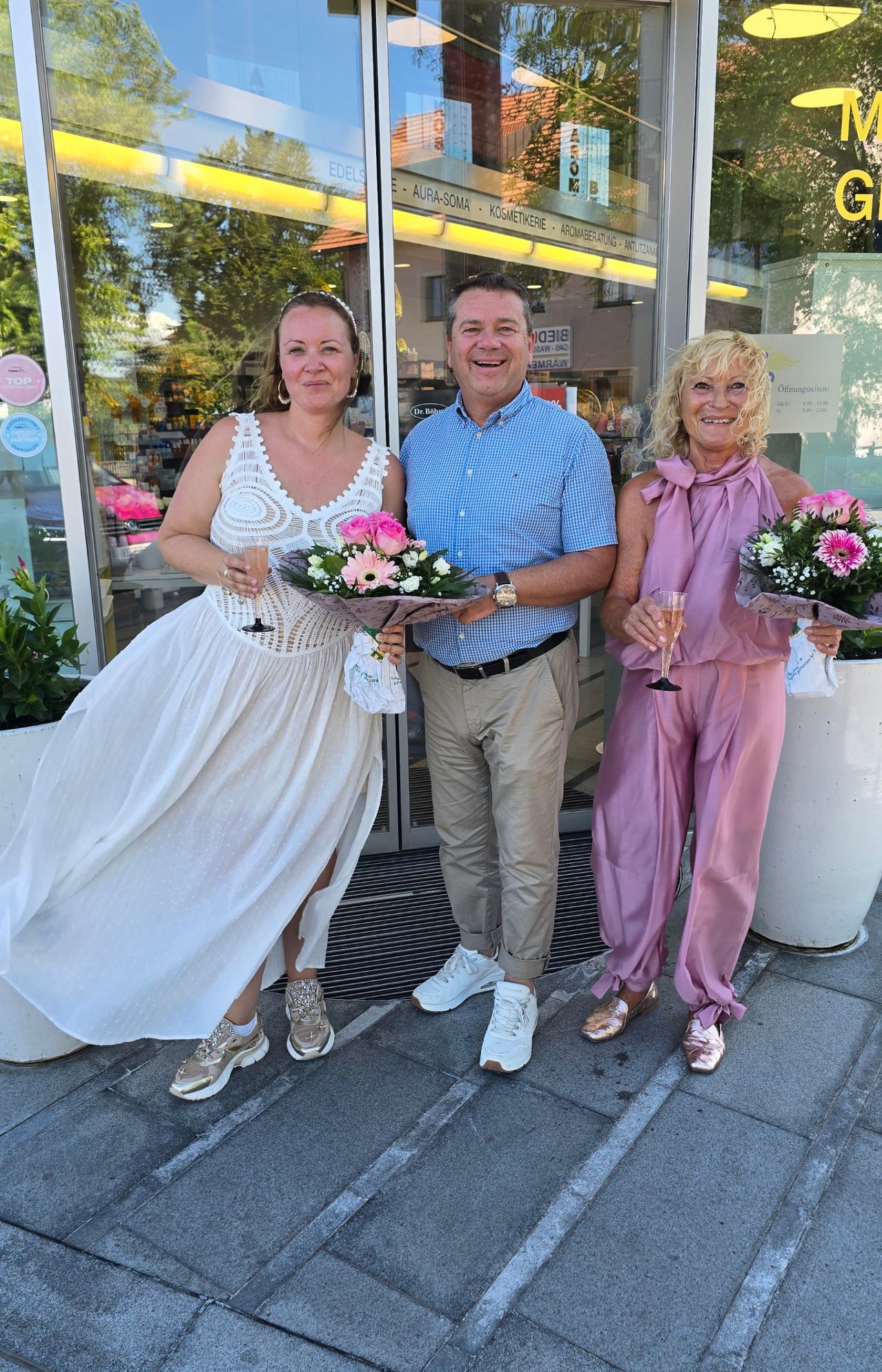 Drei Personen stehen vor einem Geschäft, zwei Frauen in Kleidern und ein Mann in einem blauen Hemd, alle halten Blumen und lächeln für ein Foto.