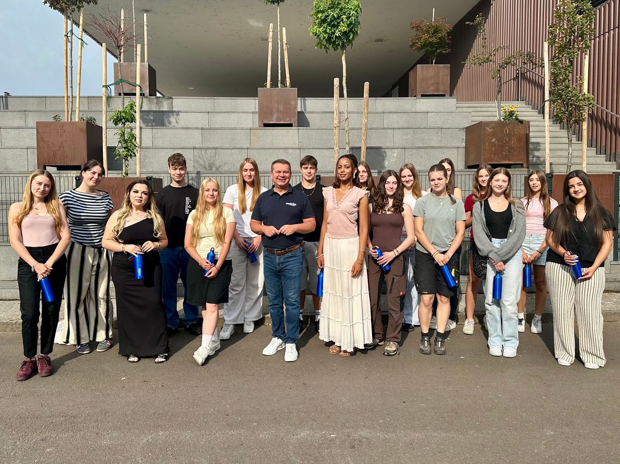Eine Gruppe junger Menschen und Erwachsene posiert für ein Foto vor einem Gebäude mit Treppen und Topfpflanzen. Sie halten blaue Wasserflaschen.