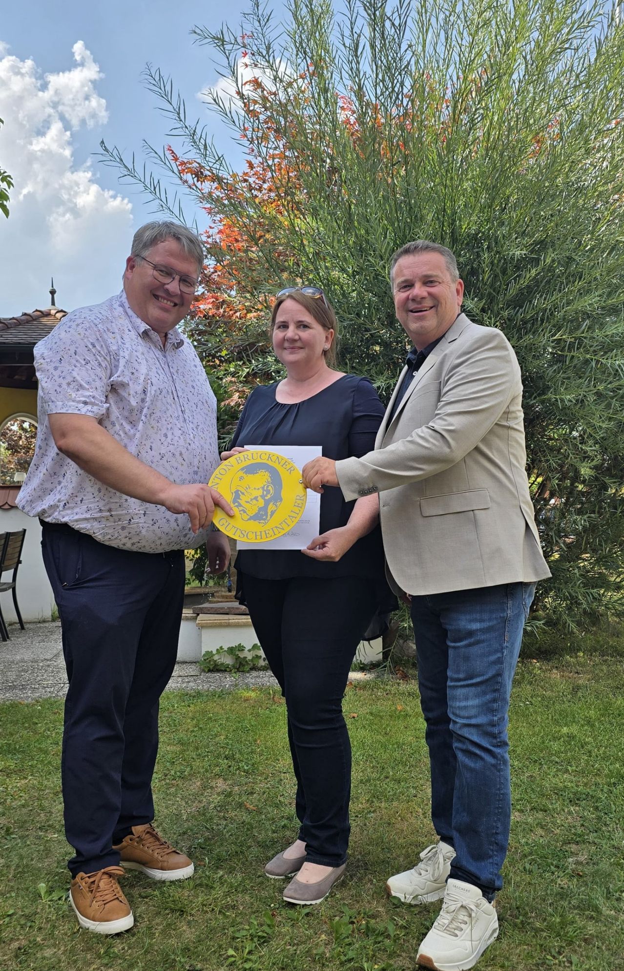 Drei Menschen stehen lächelnd in einem Garten, einer hält ein gelbes Zertifikat. Der Mann links trägt eine Brille und ein gemustertes Hemd. Die Frau in der Mitte hält ein Zertifikat mit einem Löwenlogo. Der Mann rechts trägt einen Anzug.