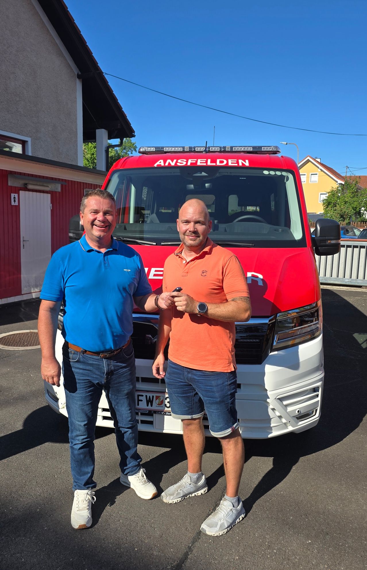 Zwei Männer stehen vor einem roten Feuerwehrauto, lächeln und posieren für ein Foto. Der Mann auf der linken Seite trägt ein blaues Hemd und Jeans, während der Mann auf der rechten Seite ein orangefarbenes Hemd und Shorts trägt.