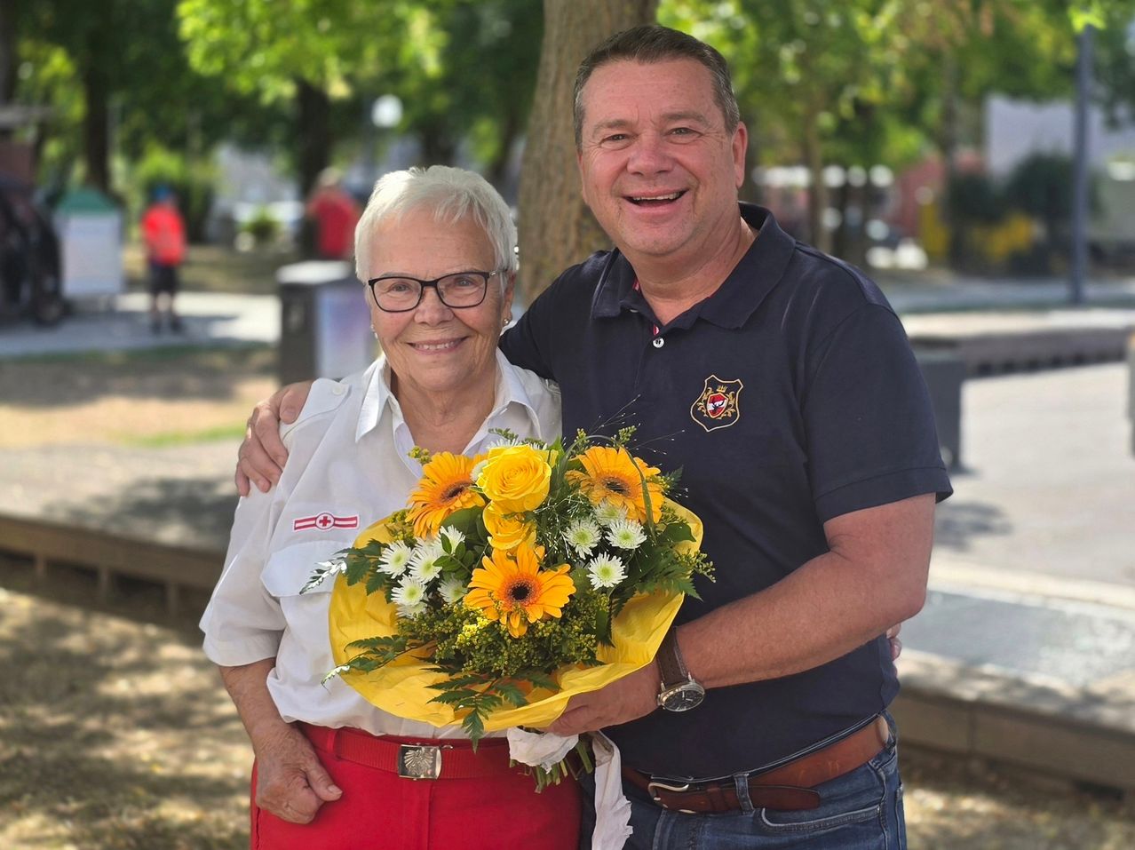 Ein Mann und eine Frau stehen lächelnd in einem Park und halten einen Blumenstrauß. Die Frau trägt eine Brille und ein weißes Hemd, während der Mann ein marineblaues Poloshirt trägt.