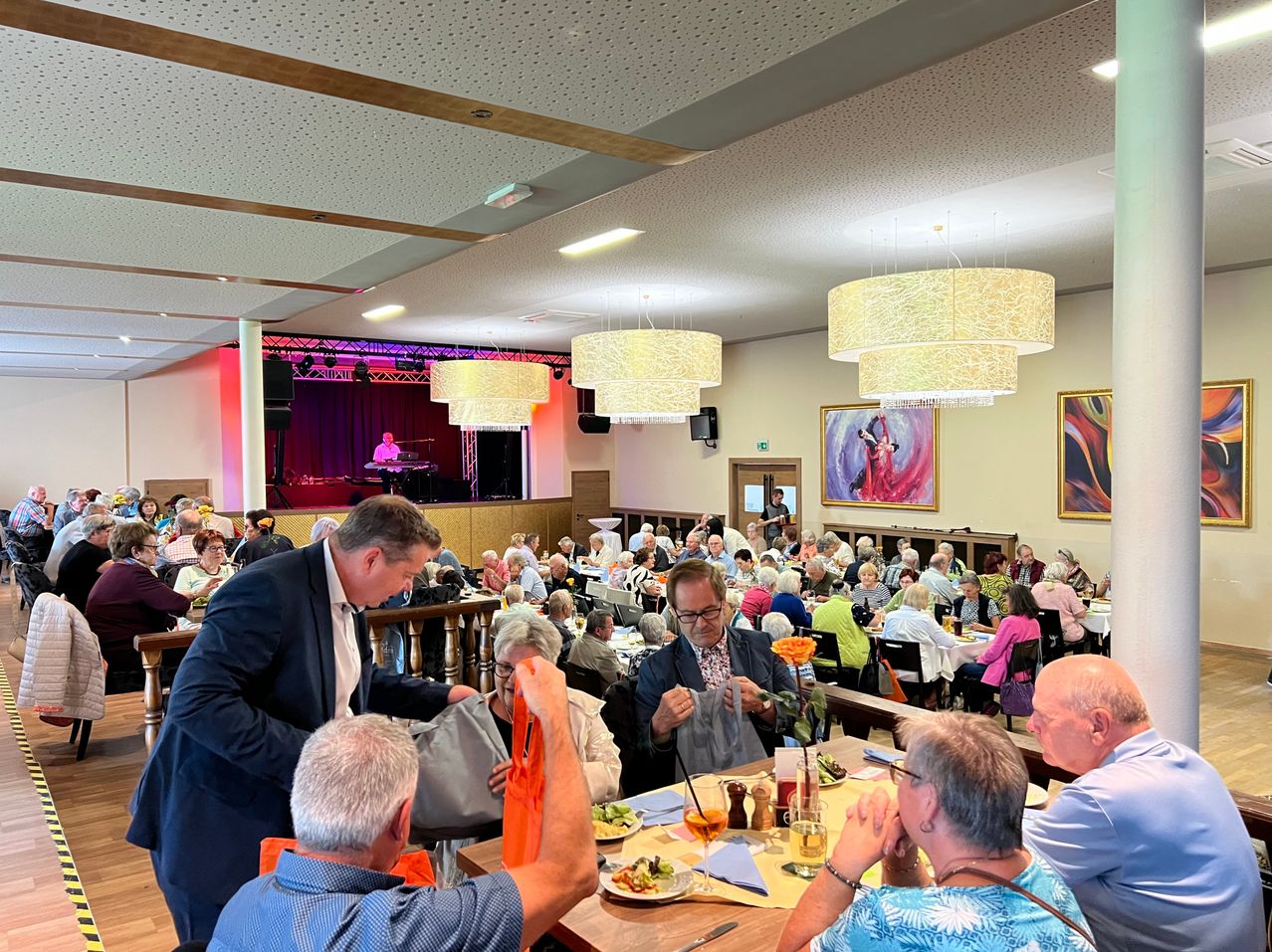 Eine Gruppe von Menschen diniert in einem Restaurant mit einer Bühne im Hintergrund, ein Mann in Anzug verteilt Taschen an die am Tisch sitzenden Personen.