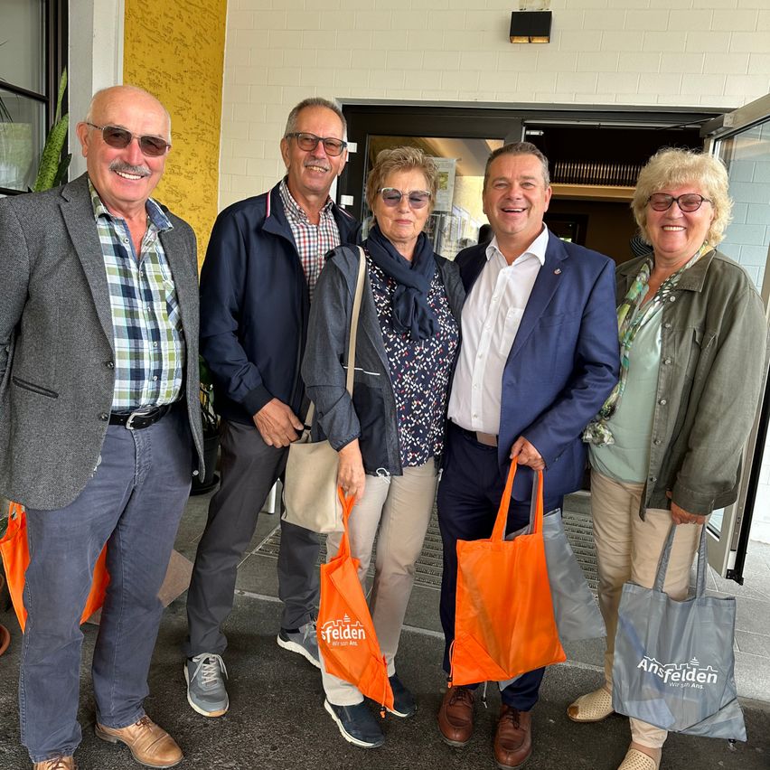 Fünf Erwachsene stehen vor einem Gebäude, lächeln und halten orange und graue Taschen. Sie sind lässig gekleidet und scheinen für ein Foto zu posieren.