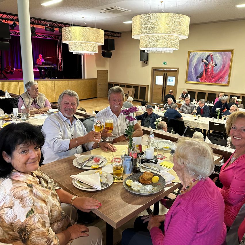 Eine Gruppe älterer Menschen sitzt in einem Restaurant um einen Tisch herum, lächelt und posiert für ein Foto. Sie halten Gläser mit Bier und Teller mit Essen. Das Restaurant hat eine Bühne mit einer Person, die Klavier spielt.