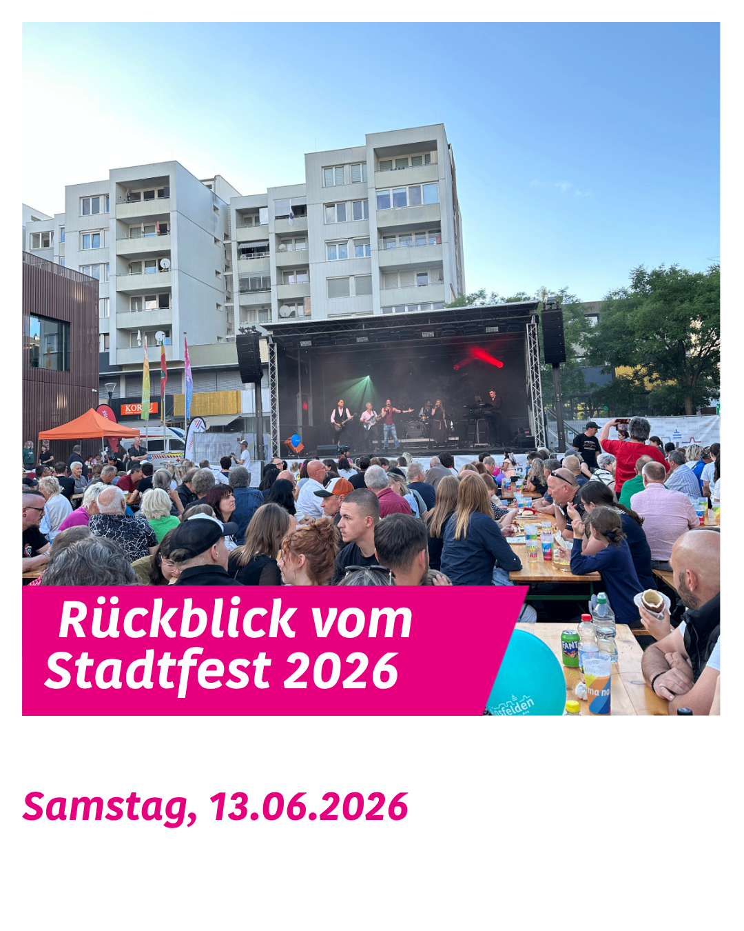 Eine Menschenmenge ist zu einem Festival versammelt und sieht einer Band auf einer Bühne mit einer großen Leinwand zu. Die Veranstaltung findet an einem sonnigen Tag statt.