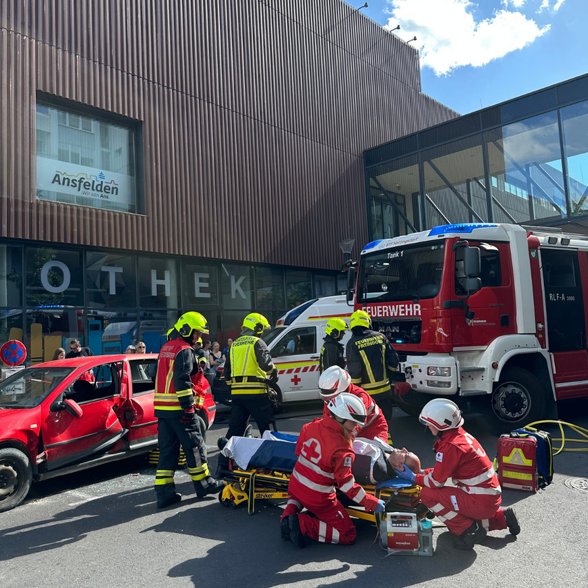 Feuerwehrleute kümmern sich um eine verletzte Person vor einem Gebäude. Ein Feuerwehrwagen und ein Krankenwagen sind in der Nähe geparkt. Die verletzte Person liegt auf einer Trage.