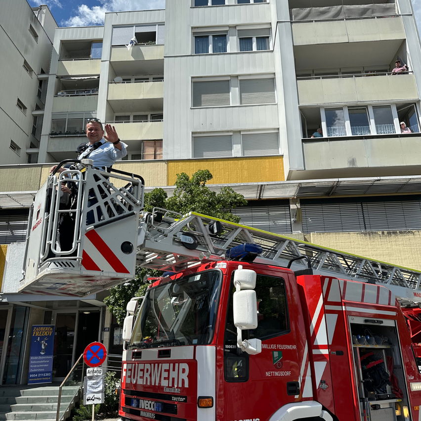 Ein Feuerwehrmann steht auf der Leiter eines roten Feuerwehrwagens vor einem Mehrfamilienhaus mit mehreren Personen auf dem Balkon.