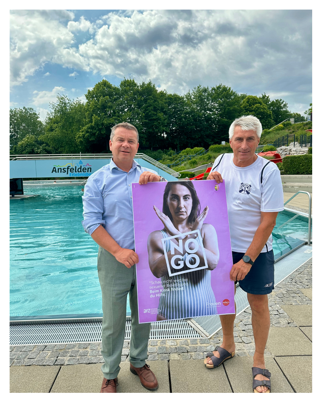 Zwei Männer stehen am Rand eines Schwimmbeckens und halten ein Plakat mit einer Frau in einer Stopp-Geste. Das Plakat trägt die Aufschrift 'No Go' und hat ein Logo unten.