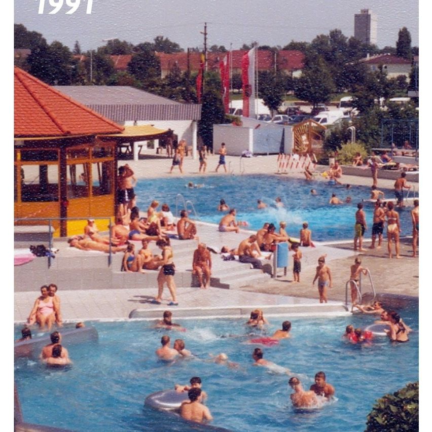 A vintage photo from 1991 shows a crowded outdoor pool area with people swimming and lounging. Flags are displayed on poles. The area is surrounded by a building with a red roof and steps leading to the pool.