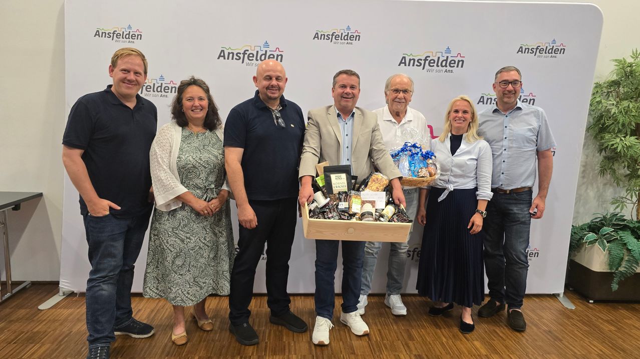 A group of people pose for a photo in front of a backdrop with the word Ansfelden. They are holding a basket filled with snacks.