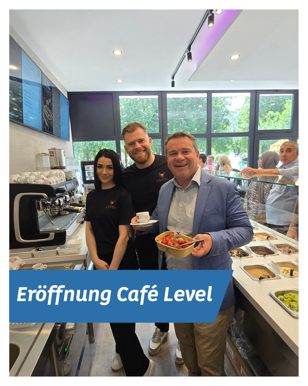 Three people, two men and a woman, stand in a cafe. They are smiling and posing for a photo. There are various foods on the counter.