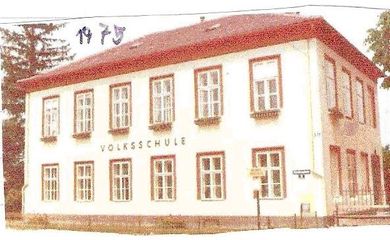 Ein altes weißes Gebäude mit einem roten Dach und vielen Fenstern, die mit 'Volksschule' beschriftet sind. Bäume sind im Hintergrund zu sehen.