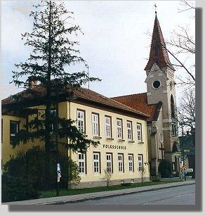 Ein altes gelbes Gebäude mit mehreren Fenstern und einem Turm, beschriftet mit 'POLESCHNICE', steht neben einer Straße mit Bäumen und einem Bürgersteig.