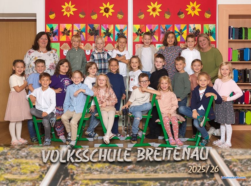 Eine Gruppe von Kindern und zwei Lehrern posiert für ein Foto vor einer mit Sonnenblumen und Herbstblättern dekorierten Wand. Die Kinder sitzen auf grünen Stühlen und lächeln. Das Foto hat einen Wasserzeichen mit dem Text 'Volksschule Brettenau 2025/26'.
