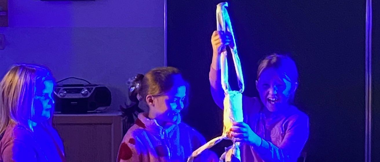 Drei junge Mädchen stehen in einem schwach beleuchteten Raum mit lila Beleuchtung und halten ein langes, eingewickeltes Objekt. Ein Mädchen lächelt, während es das Objekt hält. Hinter ihnen ist eine Uhr an der Wand angebracht.