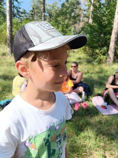 Ein kleiner Junge mit einem Schmetterling auf dem Ohr steht in einem Grasbereich, in dem andere Leute sitzen und stehen.
