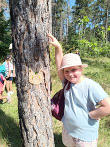 Ein junges Mädchen mit Hut steht neben einem Baum mit einer katzenförmigen Aussparung am Stamm und lächelt für ein Foto. Hinter ihr befinden sich eine Gruppe von Kindern und Erwachsenen in einem Wald.