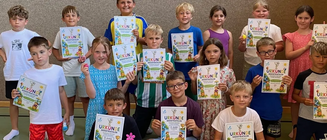 Eine Gruppe von Kindern in einer Turnhalle, die Urkunden halten und für ein Foto lächeln. Einige sitzen auf dem Boden, andere stehen, alle in Freizeitkleidung. Der Hintergrund ist eine einfache Wand mit einer schwarzen Linie auf dem Boden.