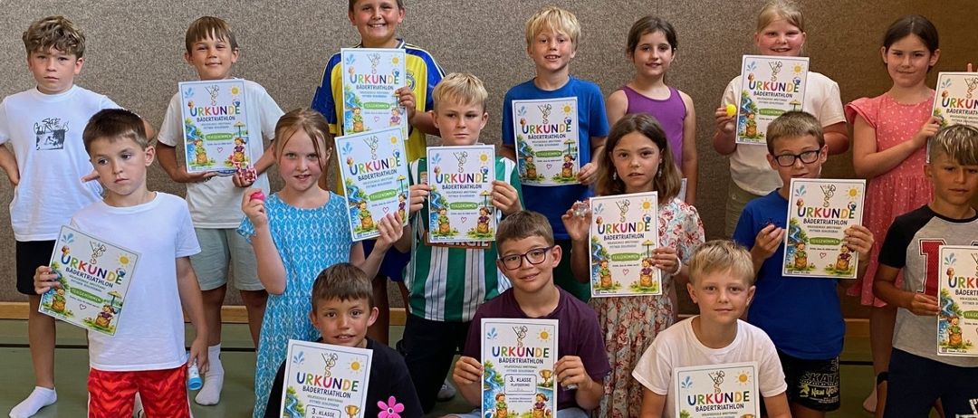 Eine Gruppe von Kindern in einer Turnhalle, die Urkunden halten und für ein Foto lächeln. Einige sitzen auf dem Boden, andere stehen, alle in Freizeitkleidung. Der Hintergrund ist eine einfache Wand mit einer schwarzen Linie auf dem Boden.