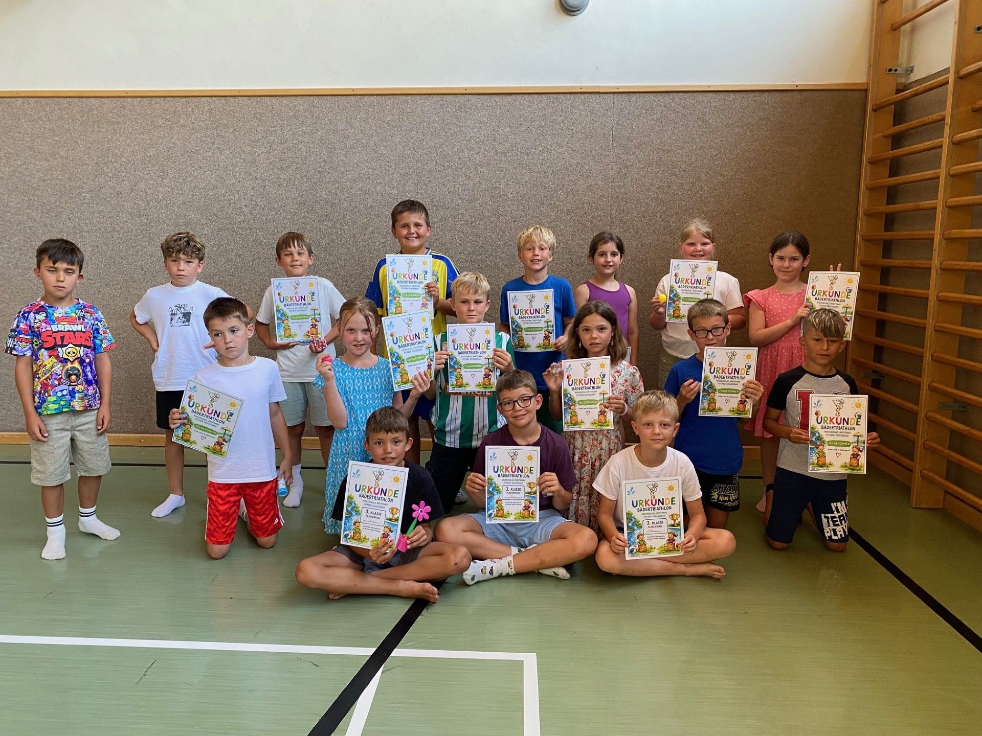 Eine Gruppe von Kindern in einer Turnhalle, die Urkunden halten. Sie lächeln und posieren für ein Foto. Einige knien, während andere stehen. Sie tragen alle Freizeitkleidung.