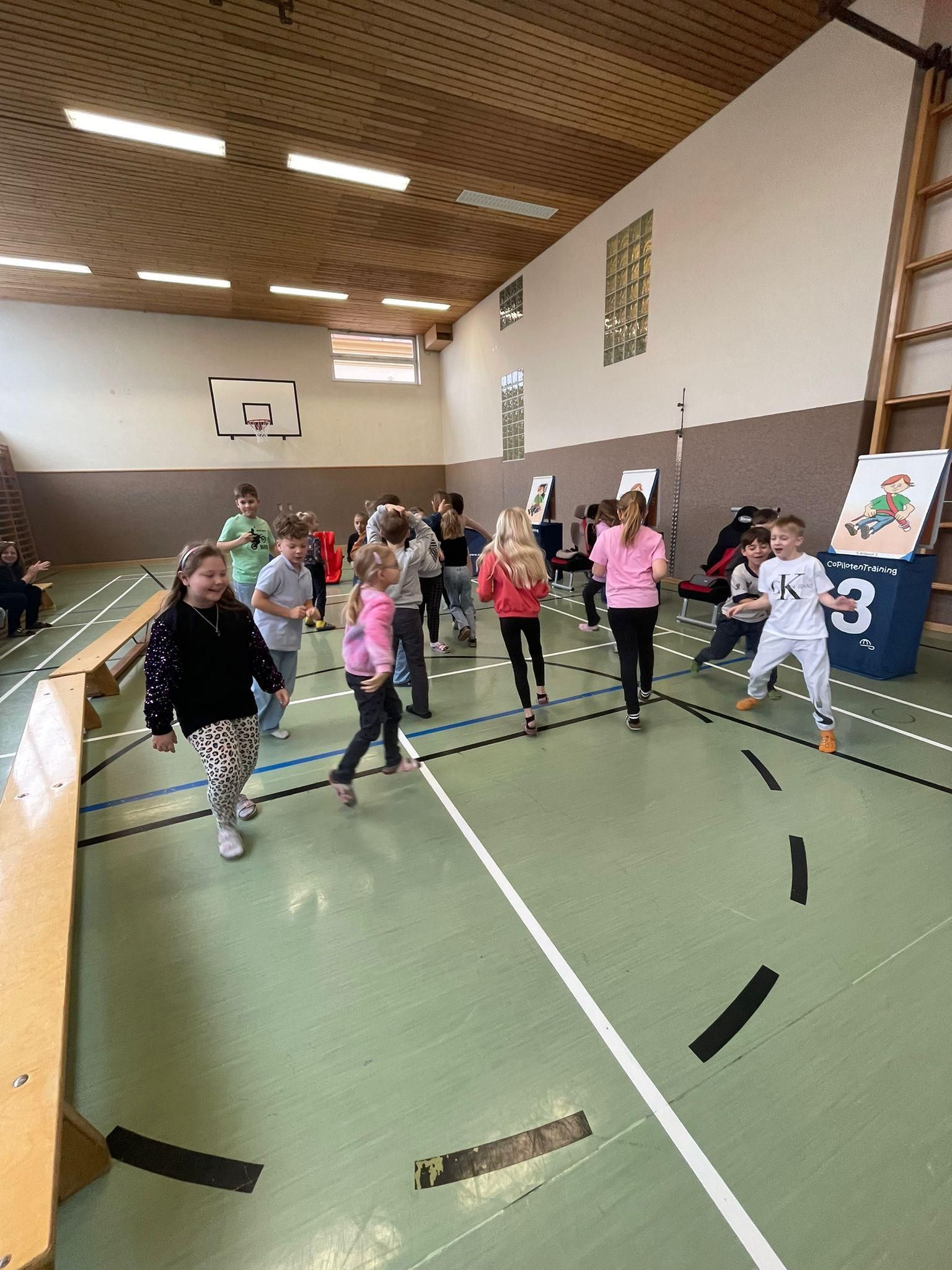 Eine Gruppe von Kindern spielt in einer Turnhalle mit einem Basketballplatz. Einige gehen, andere stehen. Es gibt Poster an der Wand und eine Leiter auf der rechten Seite.