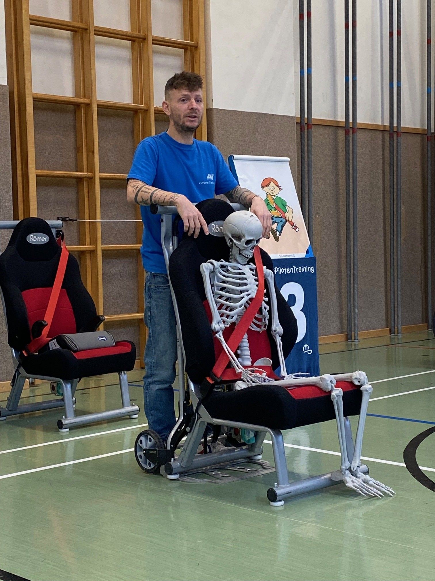 Ein Mann in einem blauen T-Shirt steht neben einem Skelett in einem Autositz und demonstriert Sicherheitsausrüstung in einer Turnhalle.