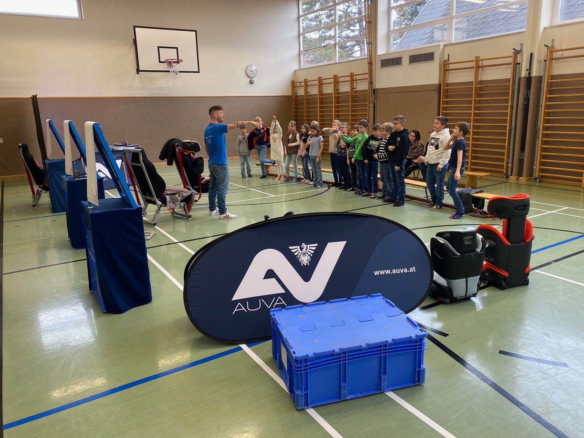 Eine Gruppe von Kindern und einem Mann stehen in einer Turnhalle. Der Mann gibt ihnen Anweisungen. Auf dem Boden befindet sich ein blaues Banner mit dem Text AUVA.