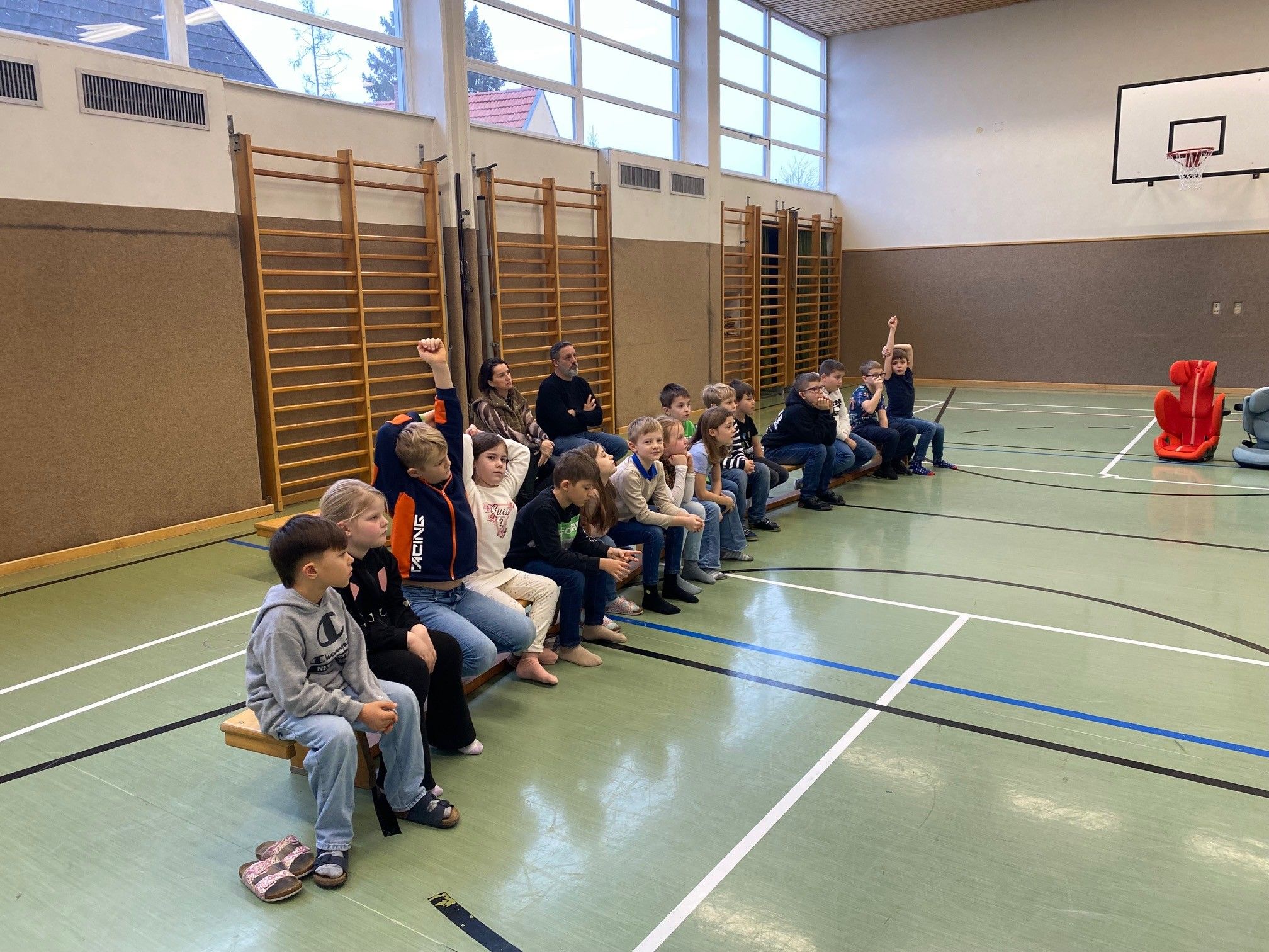 Eine Gruppe von Kindern und Erwachsenen sitzt auf einer Bank in einer Turnhalle, einige mit erhobenen Händen, während zwei Erwachsene davor stehen.