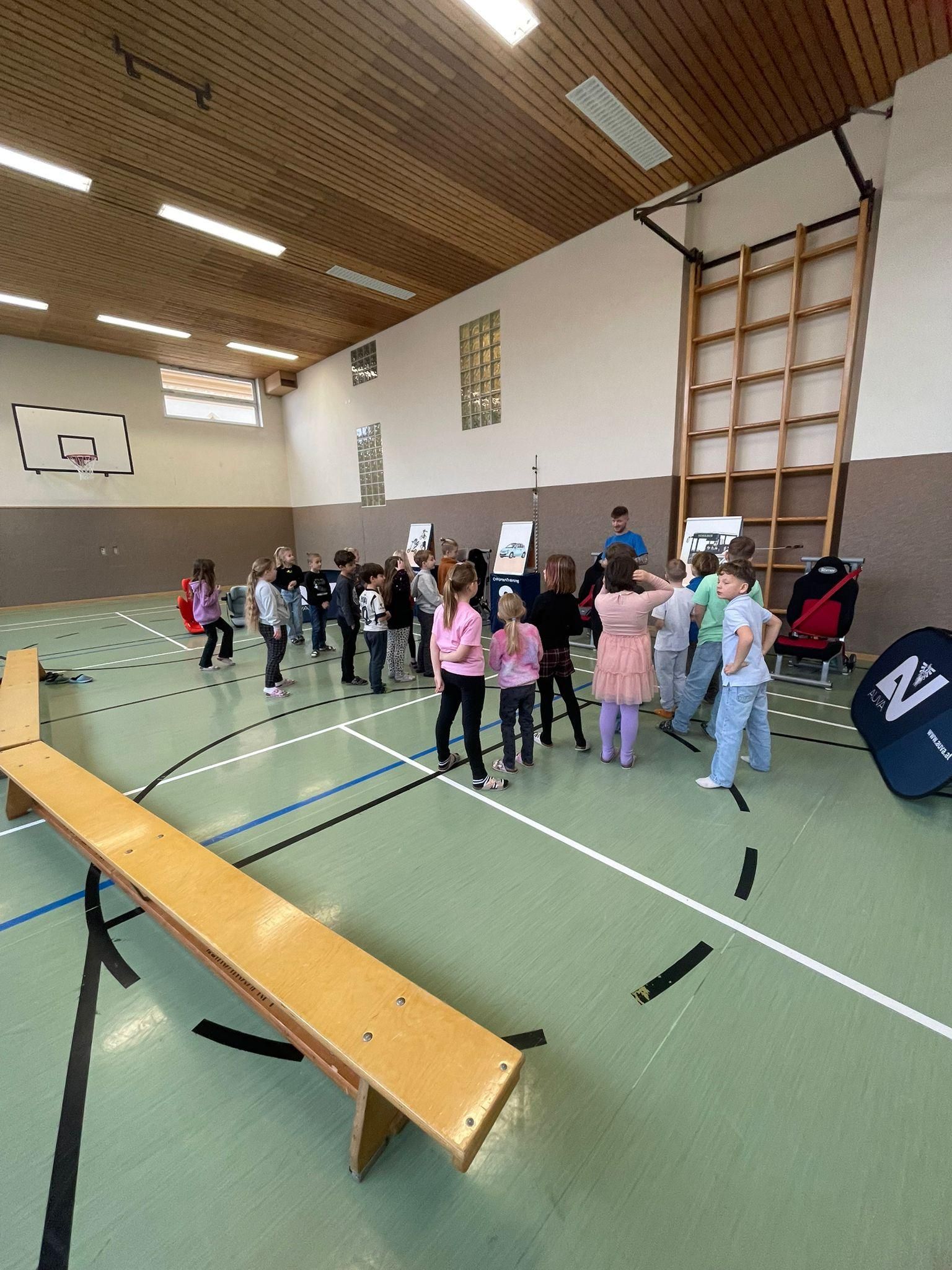 Eine Gruppe von Kindern versammelt sich in einer Turnhalle, einige stehen und andere sitzen auf einer Bank. Sie betrachten Poster an der Wand.