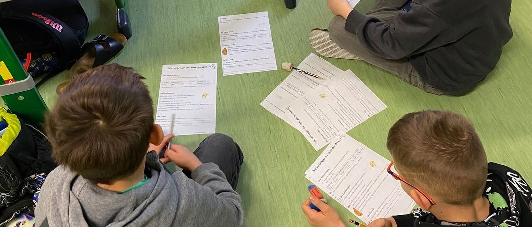 Drei Jungen sitzen auf dem Boden und beschäftigen sich mit einer Leseaktivität. Jeder Junge hält ein Stück Papier mit Text und Bildern in der Hand. Sie sind von verstreuten Papieren und einem Rucksack umgeben.