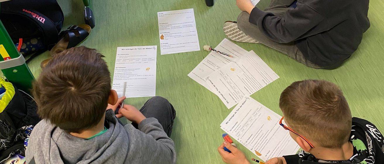Drei Jungen sitzen auf dem Boden und beschäftigen sich mit einer Leseaktivität. Jeder Junge hält ein Stück Papier mit Text und Bildern in der Hand. Sie sind von verstreuten Papieren und einem Rucksack umgeben.