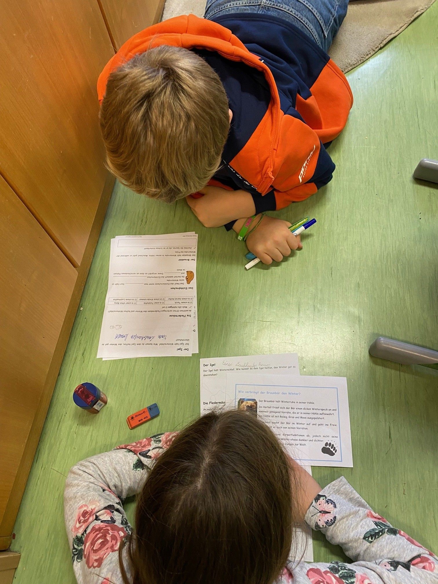 Zwei Kinder liegen auf dem Boden, eines schreibt mit einem Marker auf ein Papier, und das andere schaut auf ein Papier. Sie sind von Papieren, Markern und einem Bleistiftspitzer umgeben. Der Boden ist grün, und hinter ihnen steht ein Holzschrank.
